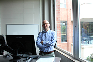 Man smiling with glasses on standing behind a desk.